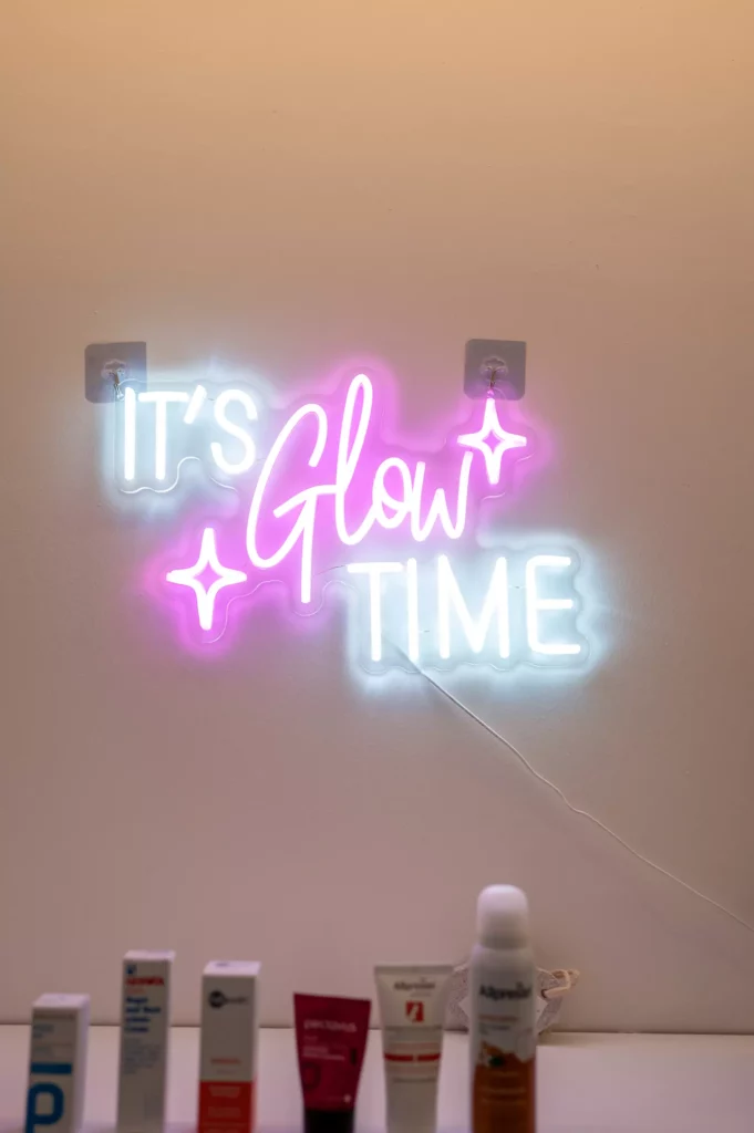 Neonlicht mit Schriftzug It's Glow Time in Weiß und Pink an Wand über Hautpflegeprodukten auf Tisch im Gesundheitszentrum