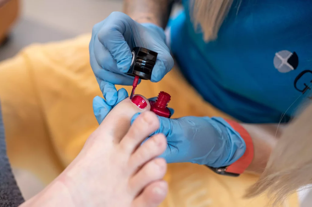 Frau mit blauen Handschuhen lackiert Zehennagel mit rotem Nagellack im Gesundheitszentrum Radstadt GHZ