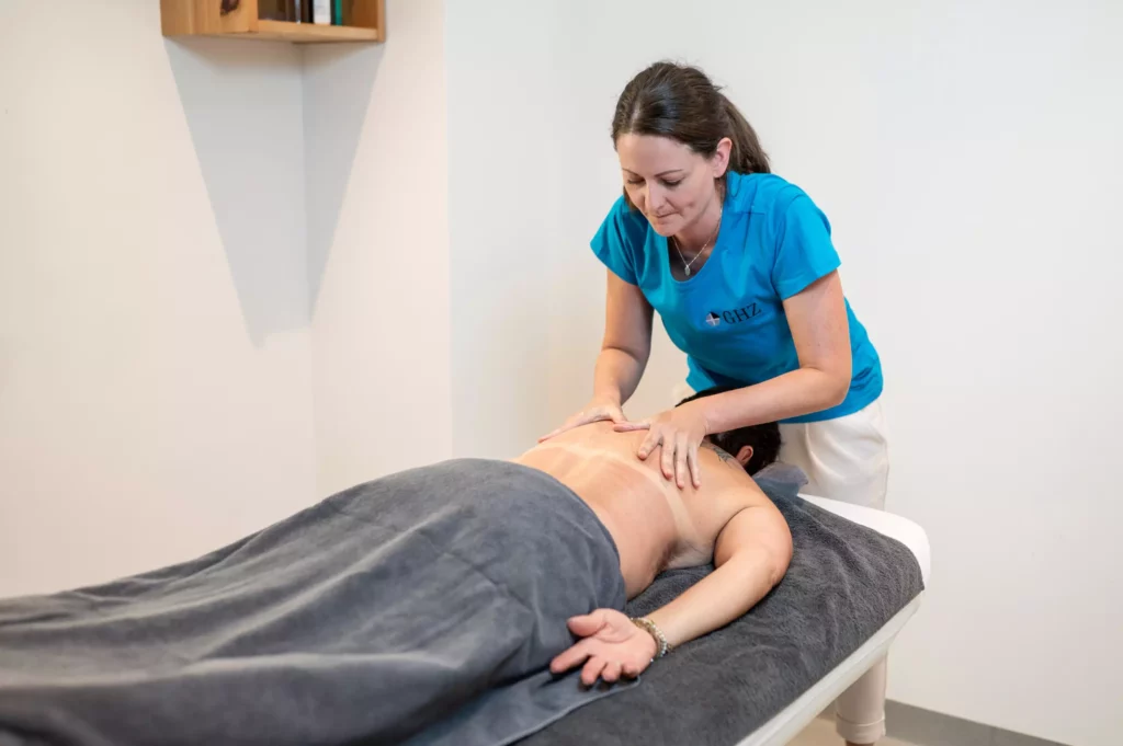 Frau in GHZ-T-Shirt massiert Rücken eines liegenden Patienten mit grauem Handtuch im Gesundheitszentrum Radstadt.