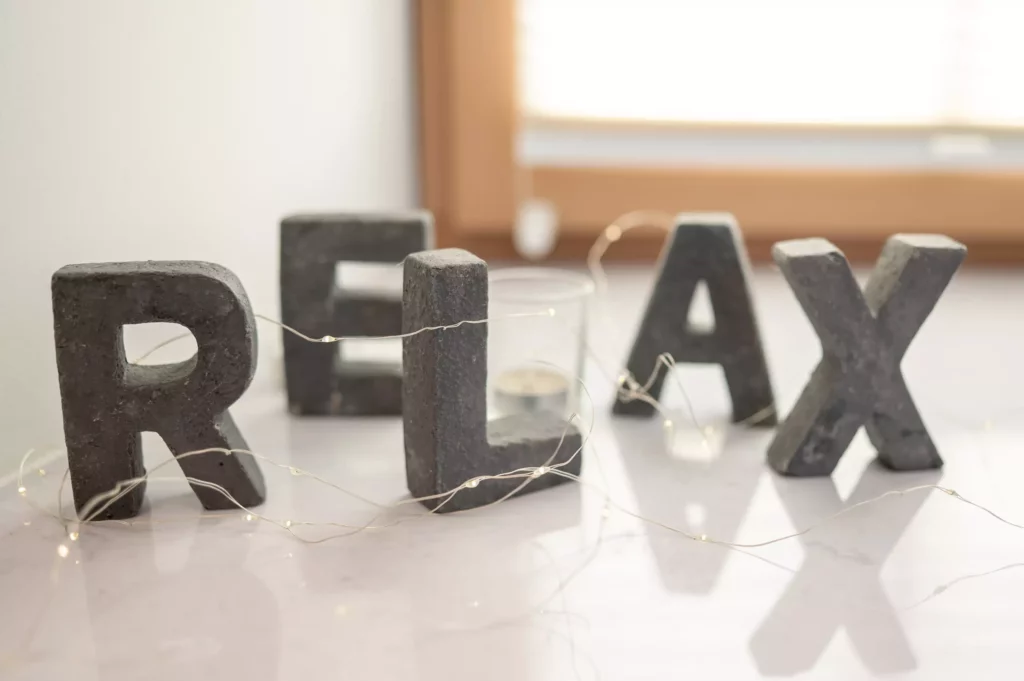 Steinerne Buchstaben bilden das Wort RELAX auf weißem Untergrund mit kleinen Lichterketten und Fenster im Hintergrund