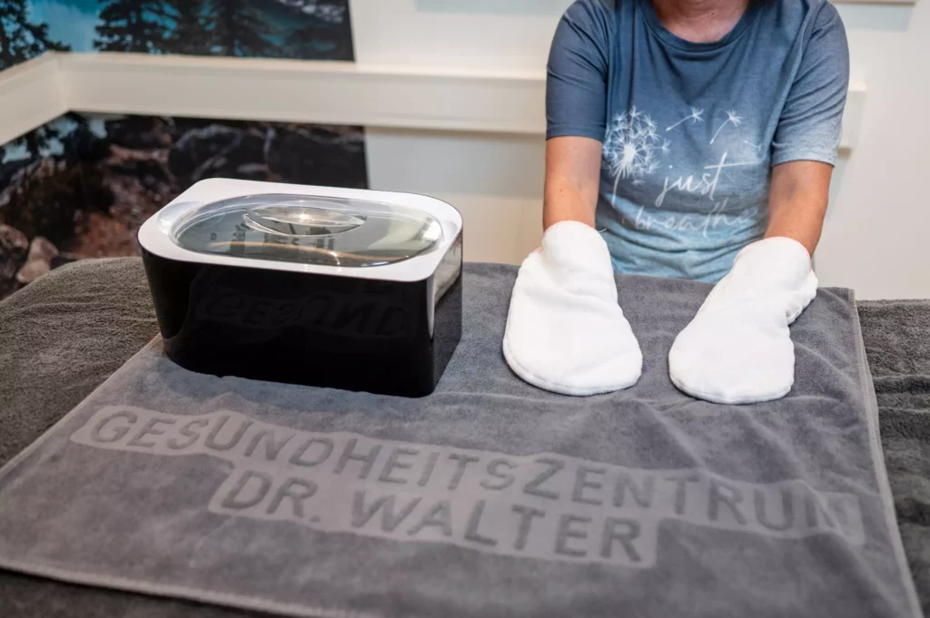 Person mit weißen Handwärmern auf einem grauen Handtuch mit Aufschrift Gesundheitszentrum Dr. Walter neben einem Wachswärmer