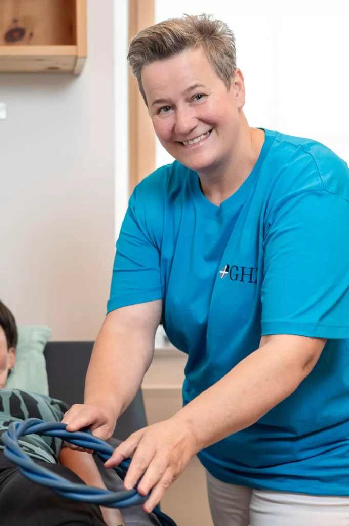 Lächelnde Therapeutin im blauen GHZ-T-Shirt hält einen blauen Trainingsring vor einer liegenden Patientin im