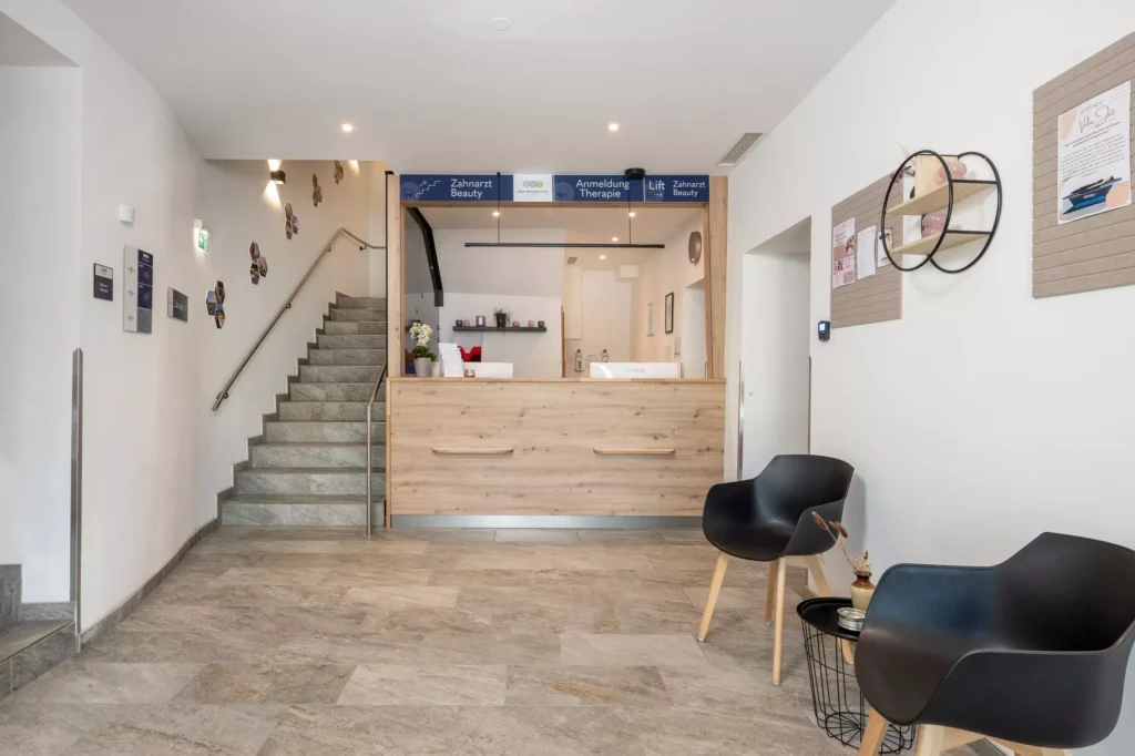 Empfangsbereich im Gesundheitszentrum Radstadt mit Holztheke, Treppe links und zwei schwarzen Stühlen rechts vor weißer Wand