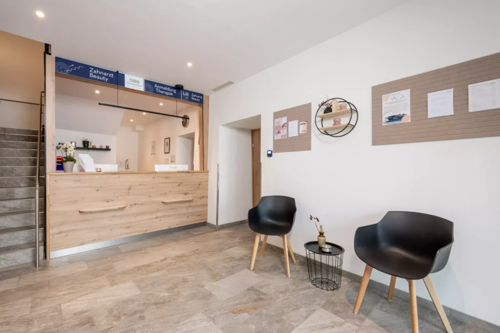 Empfangsbereich im Gesundheitszentrum Radstadt mit Holztheke, zwei schwarzen Stühlen und Informationswänden an der weißen