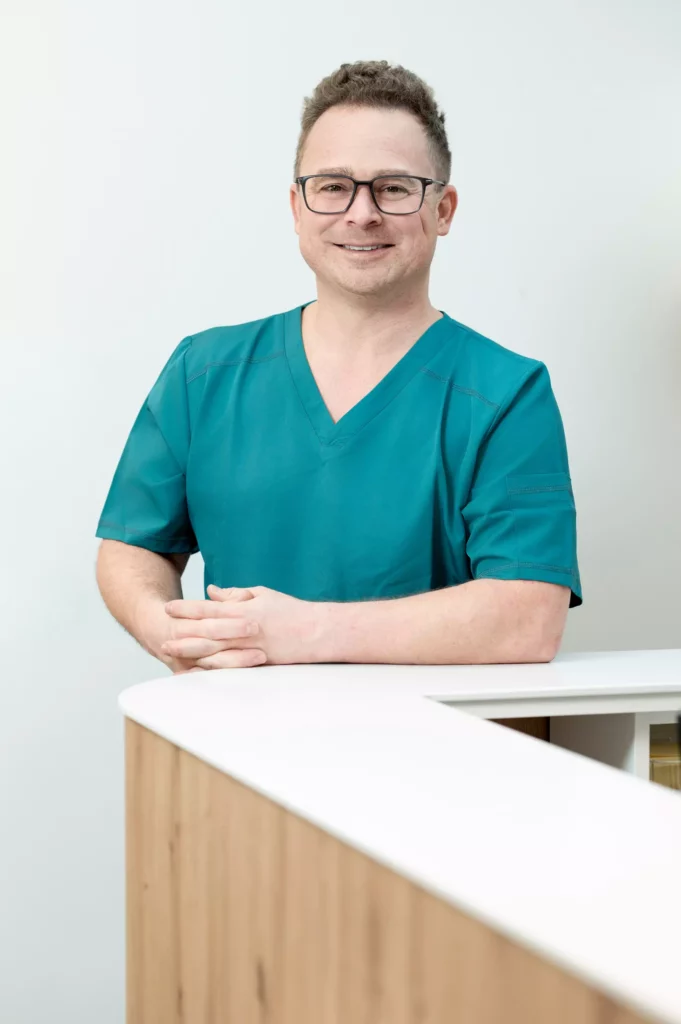 Mann in grüner medizinischer Arbeitskleidung mit Brille lächelt freundlich an einem Empfangstresen in heller Praxis